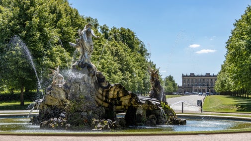 View of a large sculptural fountain in the foreground with green trees and a country house in the background with blue sky